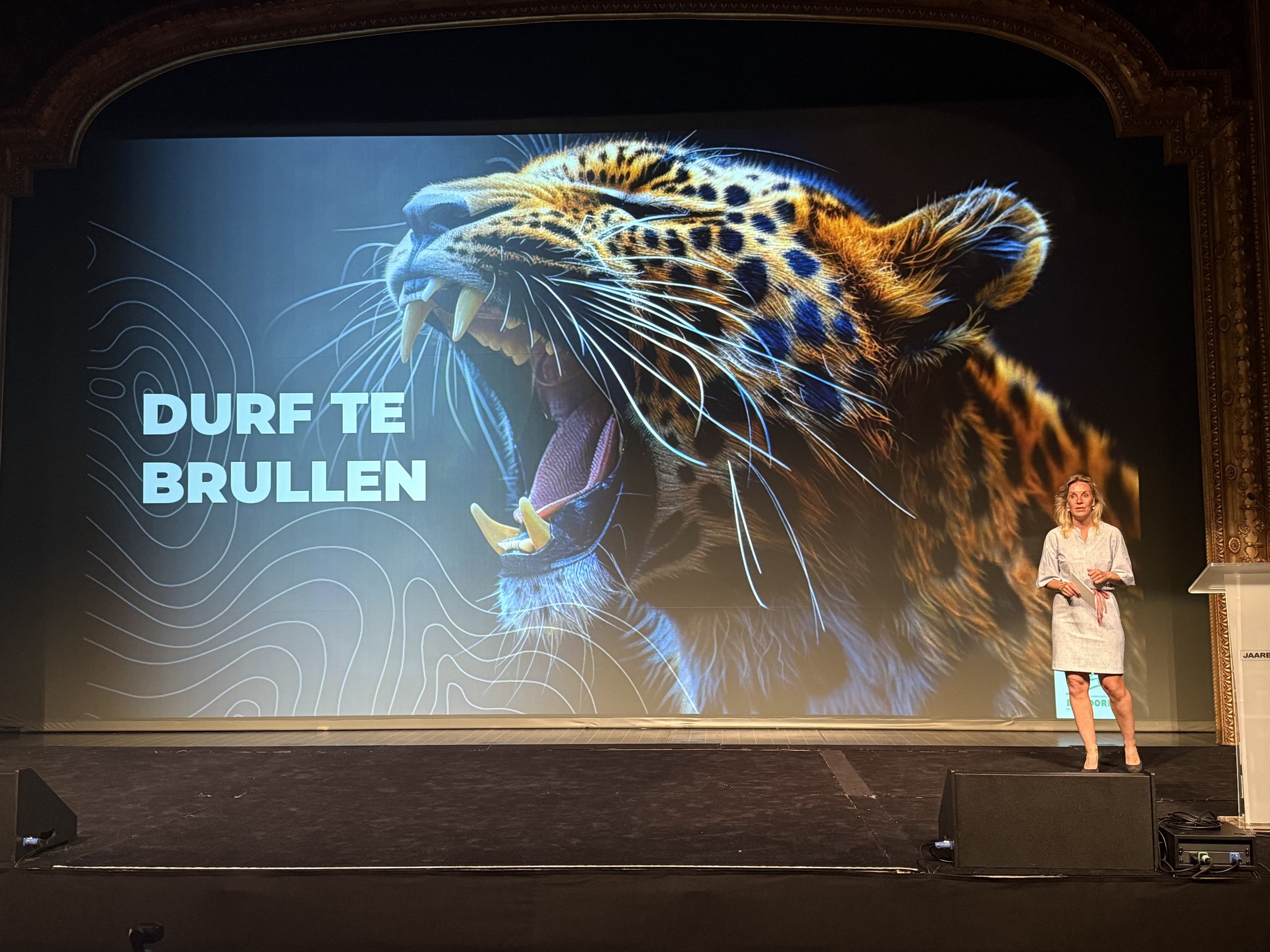 Carolien Ottjes op het podium voor een groot scherm met een brullende luipaard en de tekst Durf te brullen.